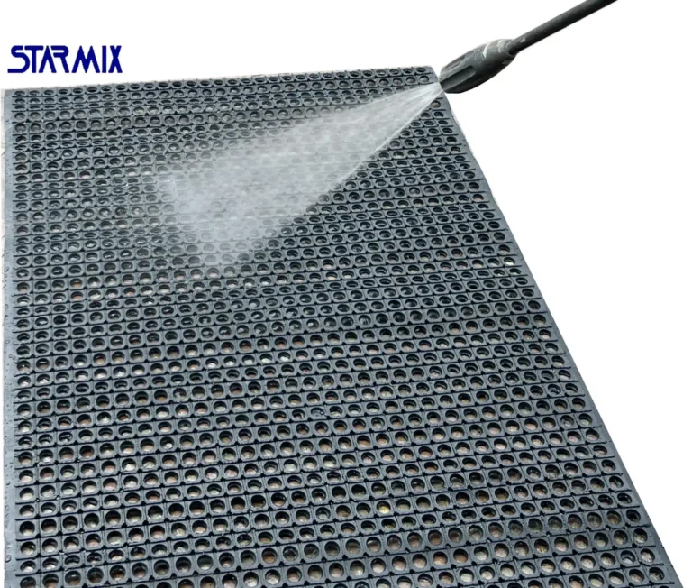 Tapete antiderrapante para cozinha industrial STARMIX sendo limpo com lavadora de alta pressão. Imagem mostra água sendo aplicada sobre o tapete de borracha nitrílica com furos.