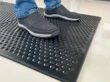 tapete de segurança antifadiga preto com superfície de domos convexos. Imagem foca na ergonomia para trabalho em pé e norma nr17.